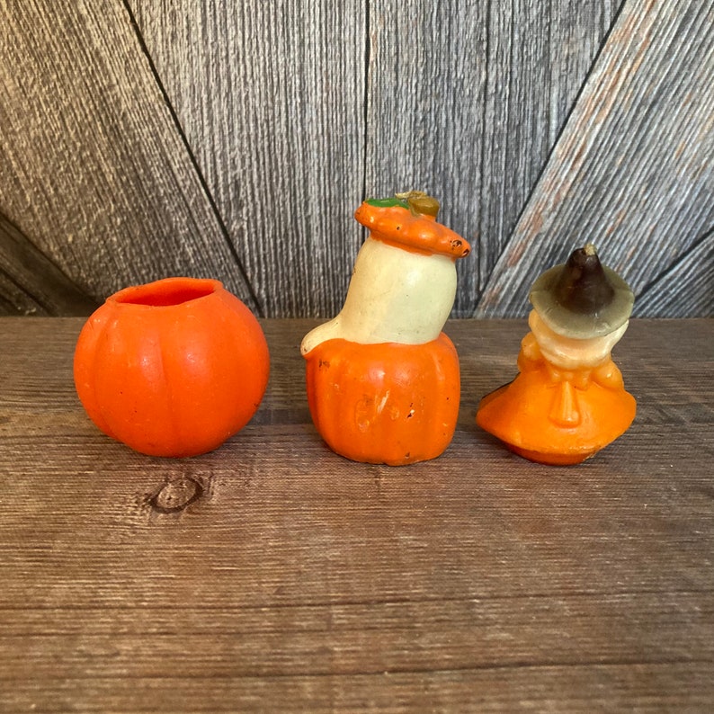 May include: Three Halloween-themed pumpkin candles. One is a plain orange pumpkin, one is a ghost with a white face and a pumpkin hat, and the last is a witch with a brown hat and orange dress.