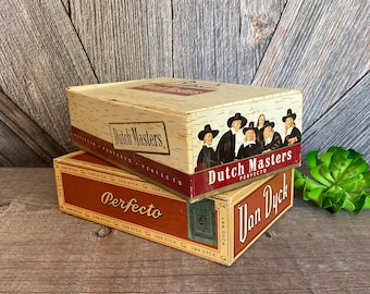 May include: Two vintage cigar boxes stacked on top of each other. The top box is brown with a wood grain pattern and says "Dutch Masters Perfecto" in gold lettering. The bottom box is red with gold lettering and says "Perfecto Van Dyck".