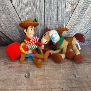 May include: Two plush toys from the movie Toy Story. One is Woody, wearing a brown cowboy hat, red scarf, blue jeans, and brown boots. The other is a plush donkey with a saddle and a green collar.
