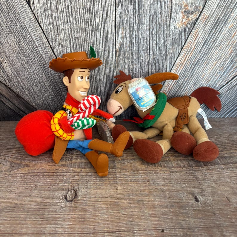 May include: Two plush toys from the movie Toy Story. One is Woody, wearing a brown cowboy hat, red scarf, blue jeans, and brown boots. The other is a plush donkey with a saddle and a green collar.