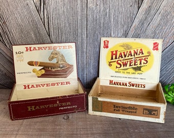 May include: Two vintage cigar boxes. The first, a burgundy box with "HARVESTER PERFECTO" text, features a cigar and horse head. The second, a cream-colored box, reads "HAVANA SWEETS" and "Invincible Foil Wrapped".