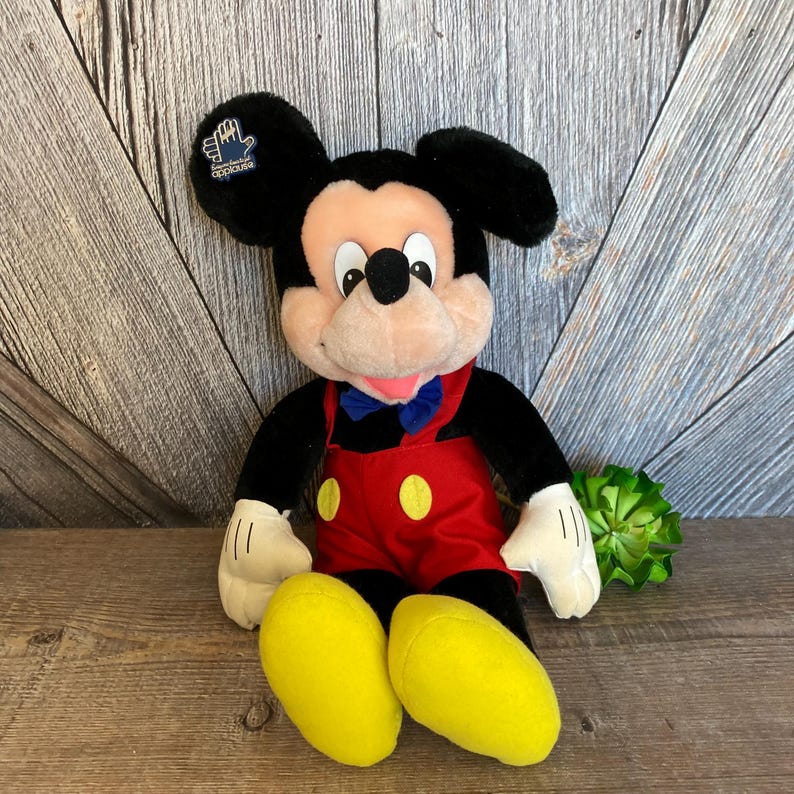 May include: A plush Mickey Mouse toy wearing red dungarees with yellow buttons and a blue bow tie. The toy has a black nose and large black ears with a "Disneyland" tag on the left ear.