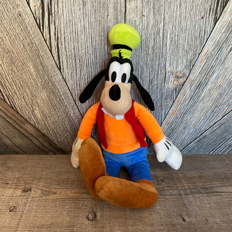 Puede incluir: Un juguete de peluche de Goofy, un personaje de dibujos animados de Disney. Lleva un chaleco rojo, pantalones vaqueros azules y un sombrero verde. Est&aacute; sentado con las piernas cruzadas y los brazos extendidos.