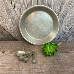 May include: A set of four silver measuring spoons on a wooden surface. The spoons are nested together and are labeled with their measurements: 1/4 tsp, 1/2 tsp, 1 tsp, and 1 tbsp. A small green succulent plant is in the background.