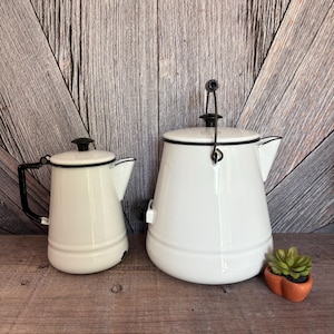 May include: Two white enamelware coffee pots with black trim and handles. The smaller pot has a black handle, while the larger pot has a black wire handle. A small orange pot with a green succulent plant sits to the right of the larger pot.