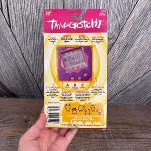 May include: A vintage Tamagotchi box in pink and yellow, featuring a purple digital pet device. The box displays instructions for feeding, lights, play, and care, with the text "Meal Snack" on the screen. The box is held by a hand.