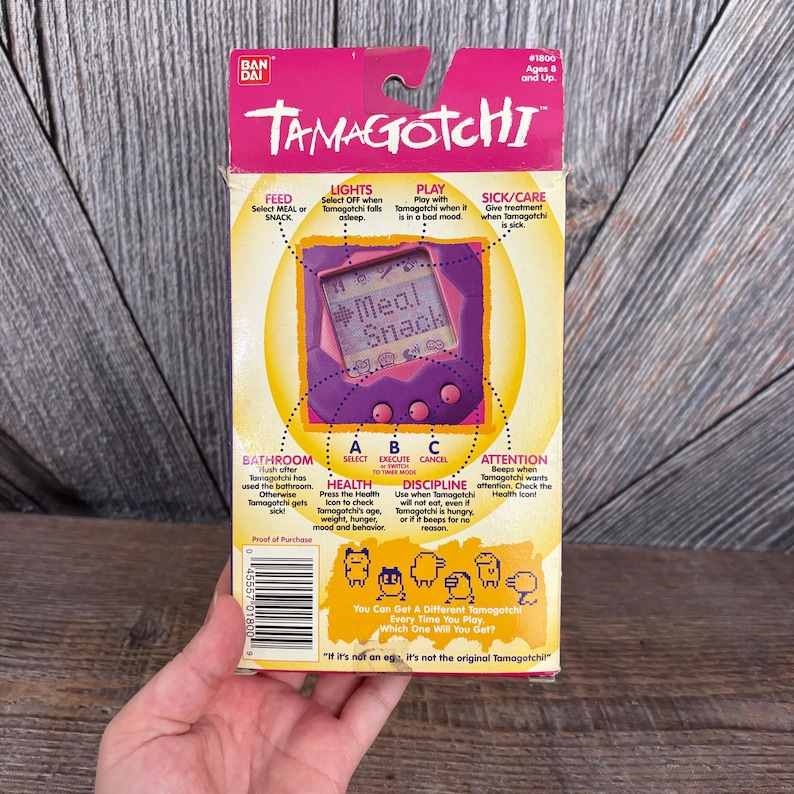May include: A vintage Tamagotchi box in pink and yellow, featuring a purple digital pet device. The box displays instructions for feeding, lights, play, and care, with the text "Meal Snack" on the screen. The box is held by a hand.