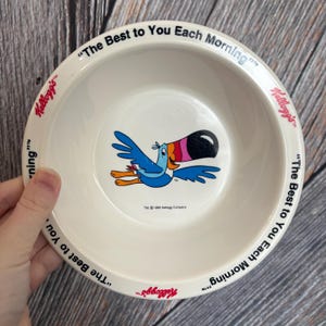 May include: A white ceramic cereal bowl featuring a cartoon toucan with a large black, pink, orange, and blue beak. The bowl has the text "The Best to You Each Morning" and the Kellogg's logo. The bowl is from 1995.