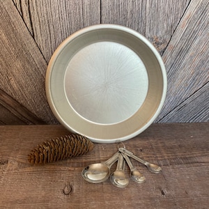 May include: A silver pie plate with a brushed finish sits on a wooden surface with a pine cone and four silver measuring spoons.