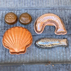 May include: Five vintage copper cake molds in the shapes of a heart, a flower, a fish, a clam shell, and a long fish. The molds are all in good condition and have a shiny copper finish.