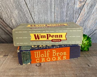 May include: Two vintage cigar boxes stacked on top of each other. The top box is green and has the text "WmPenn Perfecto" printed on it. The bottom box is blue and has the text "Wolf Bros. Crooks" printed on it.