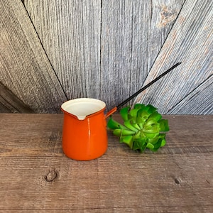May include: Orange enamelware pitcher with a black handle and white interior. The pitcher is sitting on a wooden surface.
