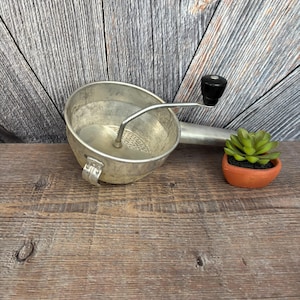 May include: A vintage, silver-toned food mill with a black handle and a perforated base. The mill is on a wooden surface next to a small terracotta pot with a green succulent plant. The background is a weathered, gray wooden wall.
