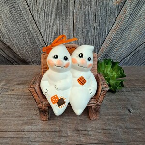 May include: Two white ceramic ghost figurines sitting on a brown wooden bench. The ghosts have orange and brown patchwork details and one has an orange bow.