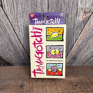 May include: A vintage Tamagotchi box with the brand name in pink, and three colourful graphic illustrations. The top of the box is purple with the brand name and age range. The box is cream-coloured and reads "the original virtual reality pet."