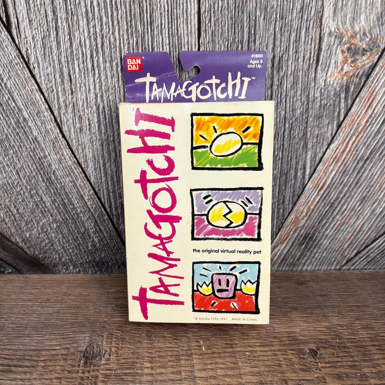 May include: A vintage Tamagotchi box with the brand name in pink, and three colourful graphic illustrations. The top of the box is purple with the brand name and age range. The box is cream-coloured and reads "the original virtual reality pet."