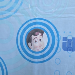 May include: Light blue fabric featuring a cartoon character's face within a blue circle. The design includes concentric blue circles and other blue shapes, creating a playful pattern. The character has brown hair and a smiling expression.