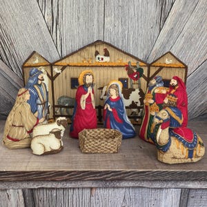 May include: A fabric nativity scene set featuring a stable backdrop with a rooster, a cow, and sheep. The figures include Joseph in red, Mary in blue, a shepherd, three wise figures, and a camel. A small woven basket sits in the center foreground.