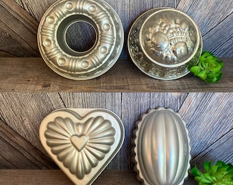 Op de afbeelding: Vier vintage zilverkleurige metalen cakevormen. De vormen zijn in de vorm van een ring, een hart, een ovaal en een rond fruitontwerp. De vormen zijn op een houten plank geplaatst.
