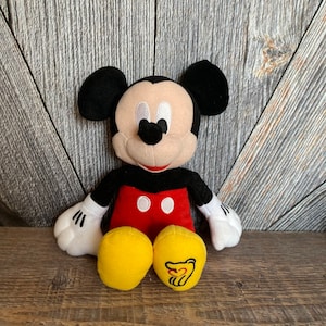 May include: A plush Mickey Mouse toy with black ears, a red shirt with two black buttons, and yellow pants. The toy is sitting on a wooden surface.