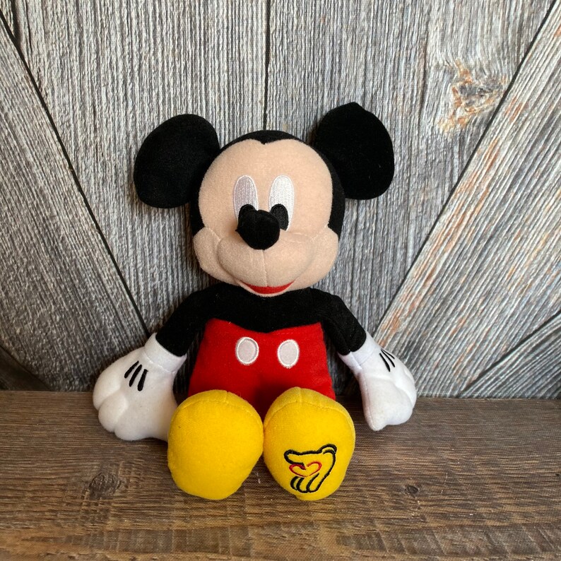 May include: A plush Mickey Mouse toy with black ears, a red shirt with two black buttons, and yellow pants. The toy is sitting on a wooden surface.