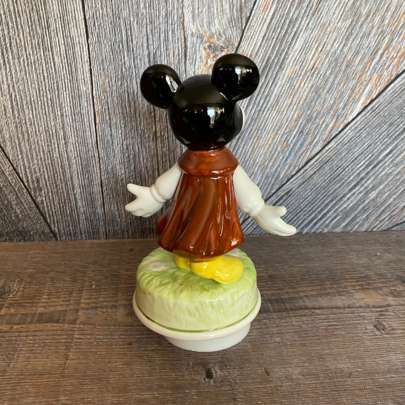 Puede incluir: Una figura de caja de m&uacute;sica de Mickey Mouse de cer&aacute;mica. Mickey lleva una chaqueta marr&oacute;n y zapatos amarillos. Est&aacute; de pie sobre una base verde con flores blancas.