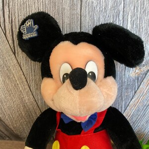May include: A plush Mickey Mouse toy wearing a red and yellow jumpsuit with a blue bow tie. The toy has a black nose and large black ears. A small blue and gold tag with the text "Applause" is attached to one of the ears.