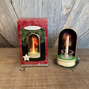May include: A white and gold Christmas ornament depicting the Washington Monument with flashing lights and sound. The ornament is in a clear dome with a green base and is surrounded by American flags. The box reads "Keepsake Ornament" and "The Washington Monument Magic".