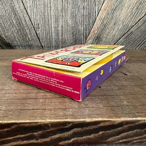 May include: A colourful Tamagotchi box with a pink, yellow, and purple design. The box features cartoon graphics and the text "Tamagotchi" in large letters. The box is open, revealing the product inside. The text "MADE IN CHINA" is visible.