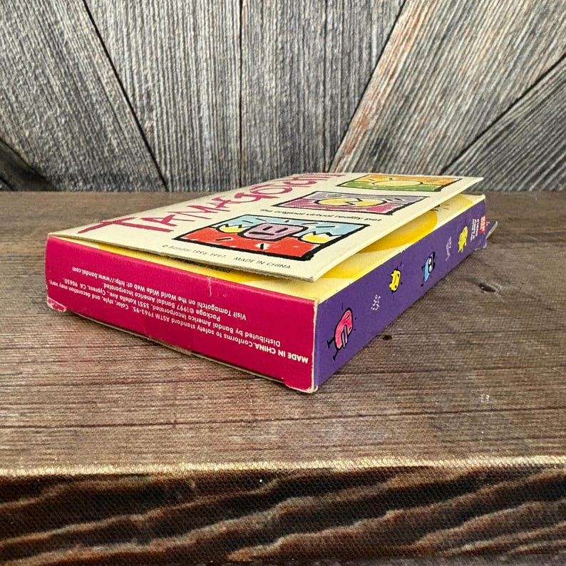 May include: A colourful Tamagotchi box with a pink, yellow, and purple design. The box features cartoon graphics and the text "Tamagotchi" in large letters. The box is open, revealing the product inside. The text "MADE IN CHINA" is visible.