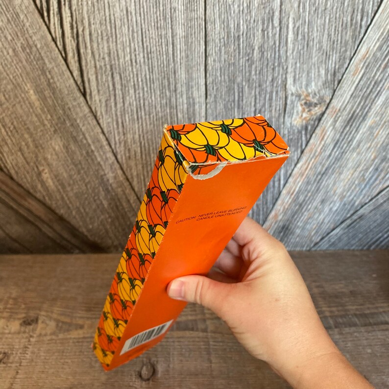 May include: An orange box with a repeating pattern of yellow and orange pumpkins. The box has the text "CAUTION: NEVER LEAVE BURNING CANDLE UNATTENDED". The box is held by a person.