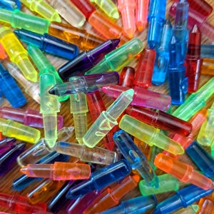 May include: A close-up shot of a pile of colorful, translucent plastic lipstick-shaped objects. The objects are in various colors, including yellow, green, blue, red, orange, and purple. The objects are scattered on a wooden surface.