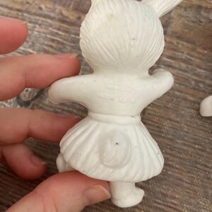 May include: White ceramic bunny figurine with a skirt and a tail. The figurine is marked "Made in Hong Kong".