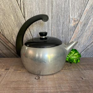 May include: A silver aluminum teapot with a black lid and handle. The teapot has a rounded body and a spout on the side.