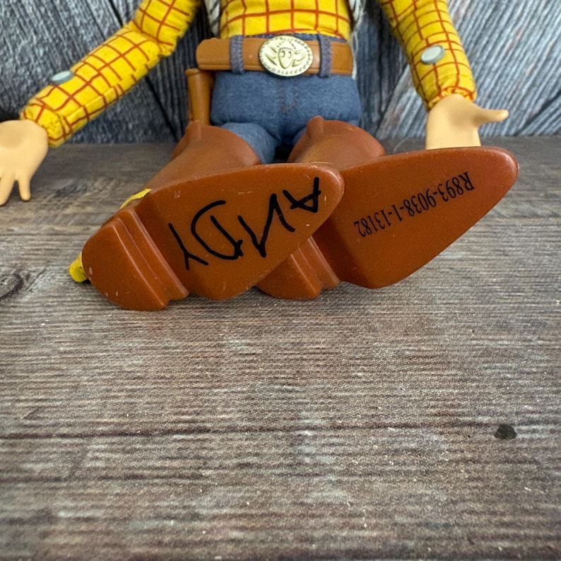 May include: A close-up of a Woody doll's brown boots with the name "ANDY" written on the soles in black marker. The doll is wearing a yellow plaid shirt, blue jeans, and a brown belt with a silver buckle.