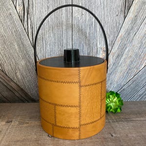 May include: A brown faux leather ice bucket with a black plastic handle and lid. The bucket has a stitched design.