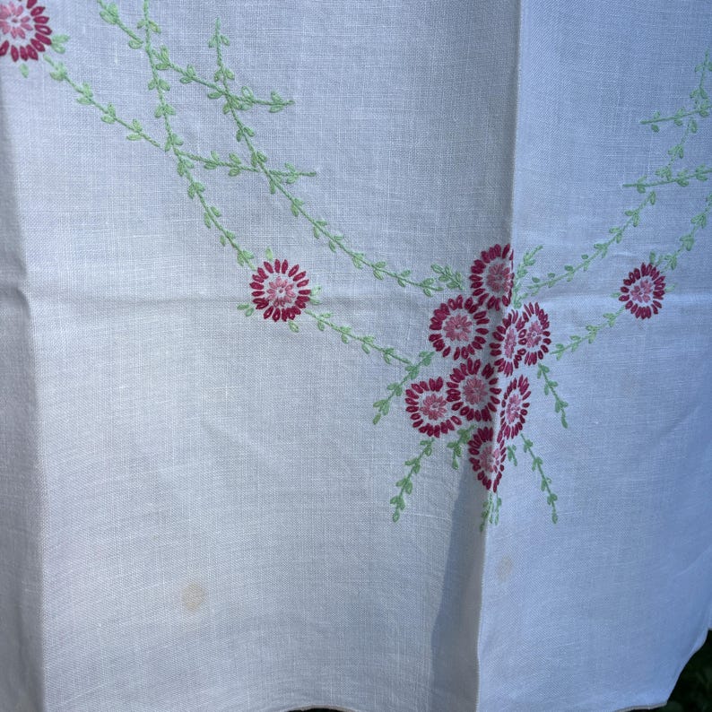 May include: White linen tablecloth with embroidered floral design. The embroidery features clusters of pink flowers and delicate green vines. The tablecloth is folded, showing the texture of the fabric and the detailed stitching.