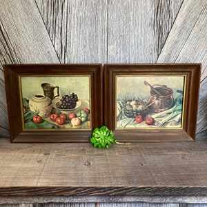 May include: Two framed still life paintings with a rustic wood background. The painting on the left features a ceramic pitcher, grapes, and fruit in a bowl. The painting on the right features a copper pot, a nest of eggs, and fruit.