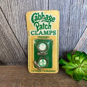 May include: A package of Cabbage Patch Kids doll clamps. The package is yellow with green text and a picture of a Cabbage Patch Kid doll. The text on the package reads "Cabbage Patch Clamps" and "I'm Unique, I'm Adoptable!"
