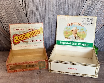 May include: Two vintage cigar boxes. The box on the left is red, white, and gold with the words "Smoke Long John Cigars Panetelas". The box on the right is white, green, and gold with the words "Optim Imported Leaf Wrapper Very Mild A Santaella y Ca - Florida Admiral".