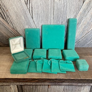 May include: A collection of 14 teal green velvet jewelry boxes and pouches. The boxes are various sizes and shapes, including square, rectangular, and round. Some boxes have a lid with the words "Eternal Gold" printed on the top.