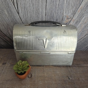 May include: Vintage metal lunchbox with a black handle and a V-shaped emblem on the front. The lunchbox has vertical ribbed detailing and a clasp closure. A small terracotta pot with a green succulent plant sits in front of the lunchbox.