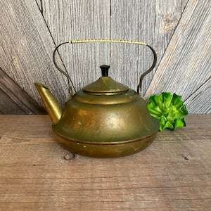 May include: A vintage brass teapot with a black knob on the lid and a white beaded handle. The teapot is sitting on a wooden surface.