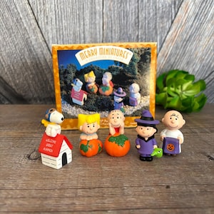 May include: A collection of Peanuts character figurines, including Snoopy, Charlie Brown, and Lucy, in Halloween costumes. The figurines are displayed in front of a box labeled "Merry Miniatures Collection". The figurines are approximately 1-2 inches tall.