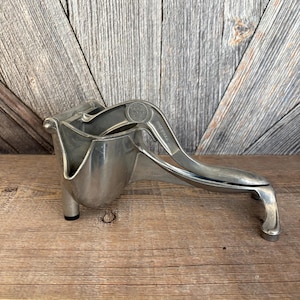 May include: A vintage, silver-colored citrus juicer. The juicer has a cup-shaped receptacle, a hinged lever, and a handle. The metal juicer is designed to extract juice from lemons, limes, and oranges. The juicer is made of metal and sits on a wooden surface.