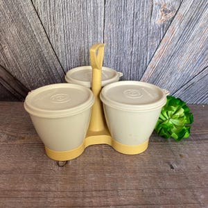 May include: A set of three beige plastic containers with lids and a yellow handle. The containers are arranged in a circle on a yellow base.