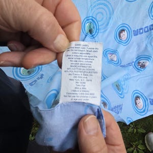 May include: Light blue twin flat sheet with a repeating pattern of cartoon characters and the word "Woody". The care label indicates the sheet is 66in x 96in (168cm x 244cm) and made of cotton and polyester.