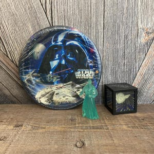 May include: A blue and white paper plate with a Star Wars theme featuring Darth Vader, a spaceship, and the text "Star Wars". A green plastic Yoda figurine and a black cube with a Yoda image inside are also pictured.