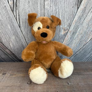May include: A brown plush dog toy with a white patch over one eye and cream-colored paws. The dog has a black nose, a pink tongue, and floppy ears. The toy is sitting on a wooden surface.