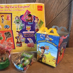 Pode incluir: Uma caixa de Happy Meal do McDonald's com o Woody do filme Toy Story 2. A caixa &eacute; amarela e vermelha com uma imagem do Woody na frente. O texto "McDonald's Happy Meal" est&aacute; impresso em branco sobre um fundo vermelho. O texto "Disney Pixar Toy Story 2" est&aacute; impresso em branco sobre um fundo amarelo. A caixa est&aacute; sobre uma mesa de madeira.